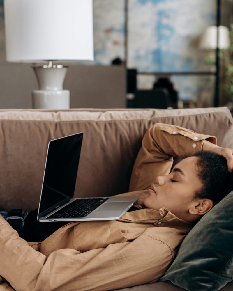 Exhausted woman rests with laptop on sofa, illustrating work-from-home fatigue.