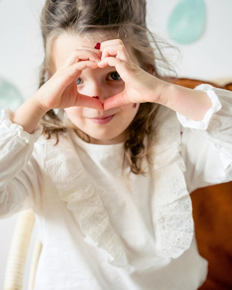 Adorable preschool girl with stickers on face looking at camera while making heart shaped gesture with hands near wall with decorative eggs during Easter celebration